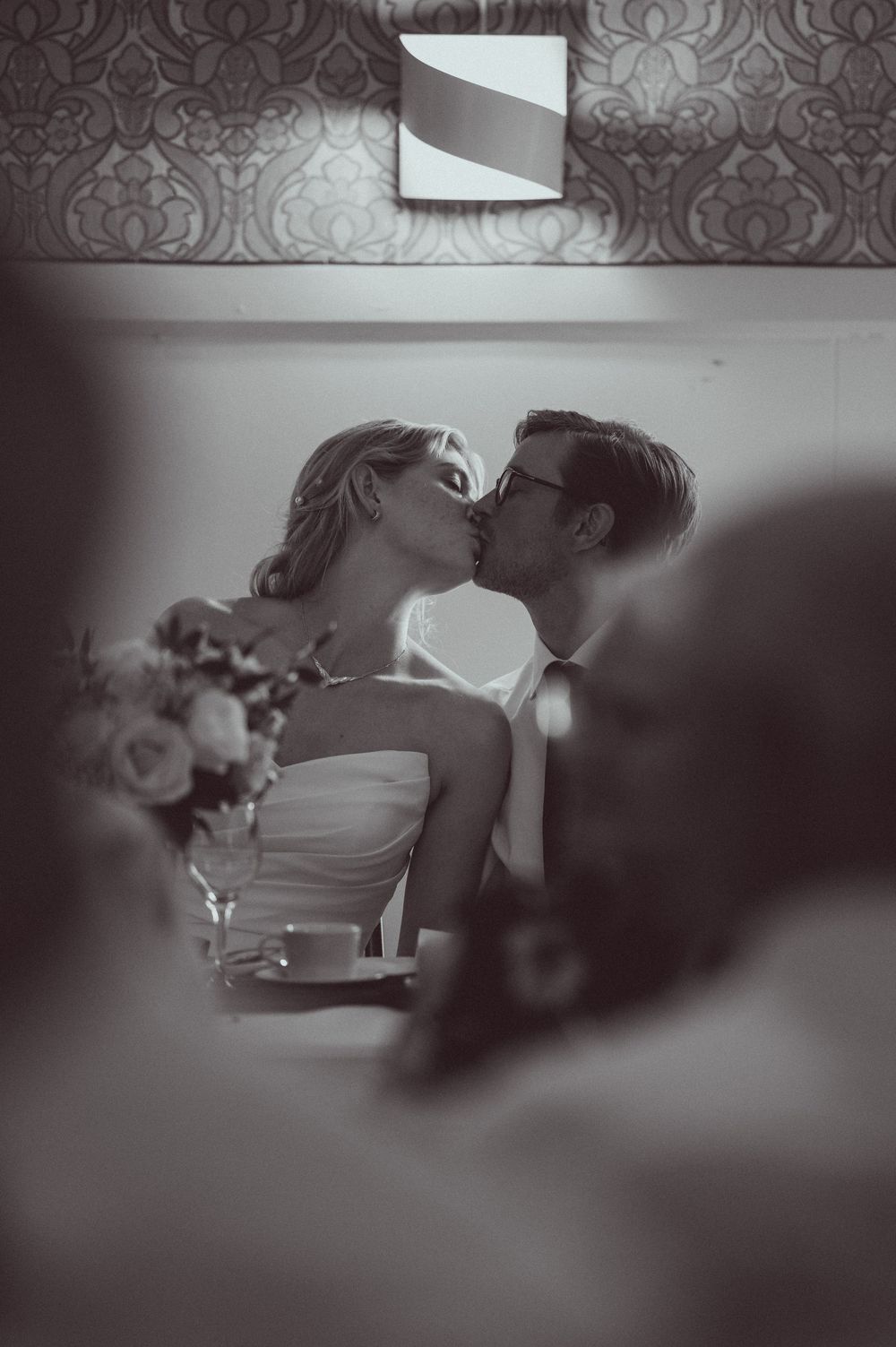 Black and white photo of a bride and groom sharing a kiss at their wedding reception table with flowers nearby.