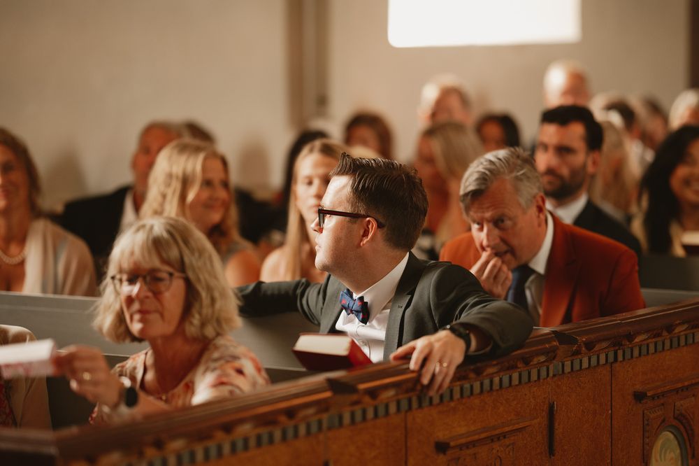 Guests seated in wooden church pews during a wedding ceremony, with a young man in a bow tie looking back.