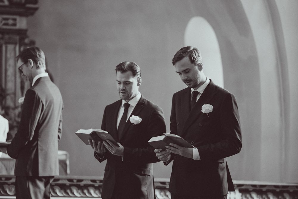 Three groomsmen in dark suits reading from books during a wedding ceremony inside a church.