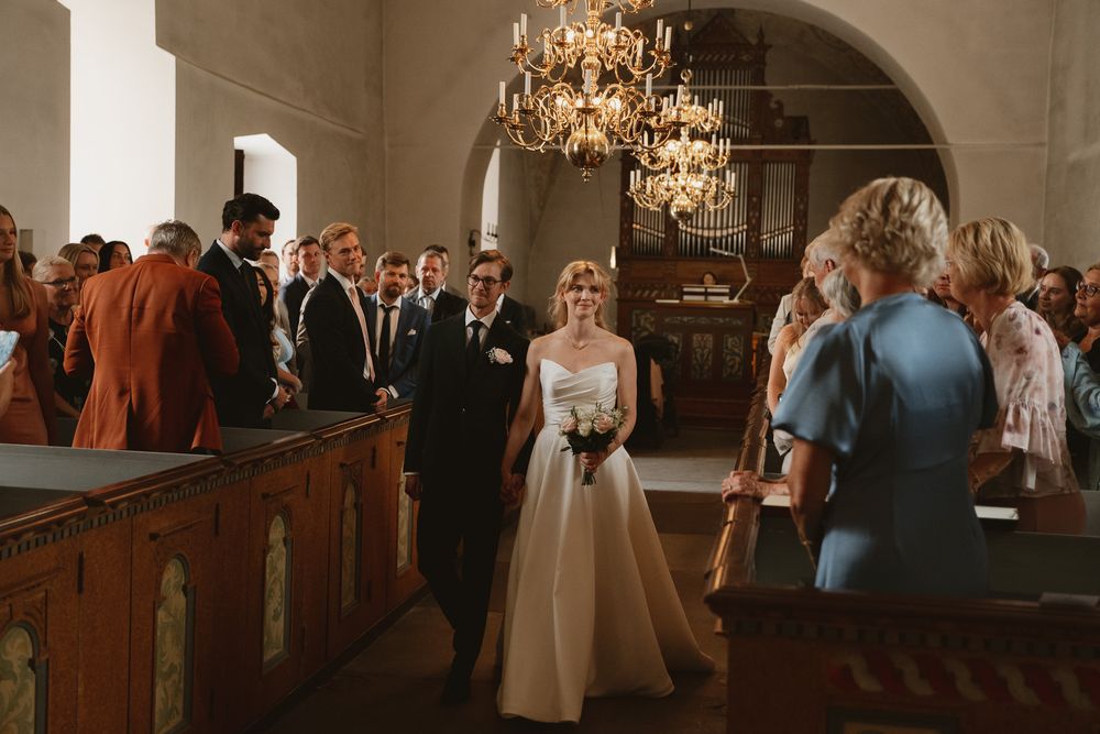 Bride walks down church aisle in elegant gown, escorted by groom, guests watching under golden chandeliers.