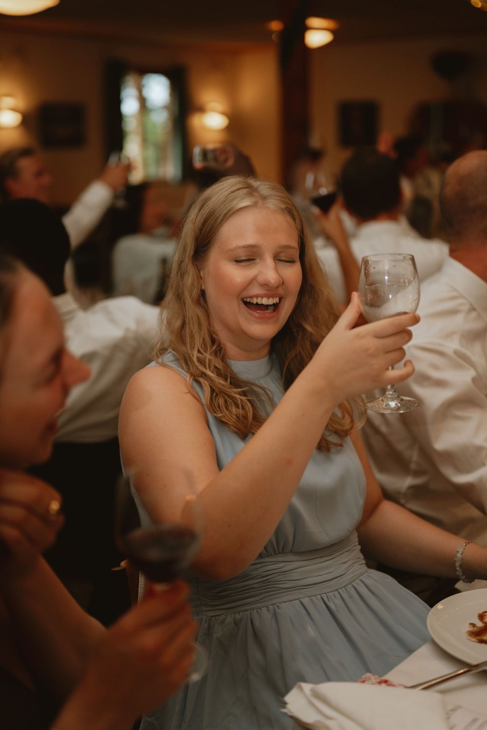 A laughing bridesmaid in a blue dress raises a champagne glass during a wedding reception toast.