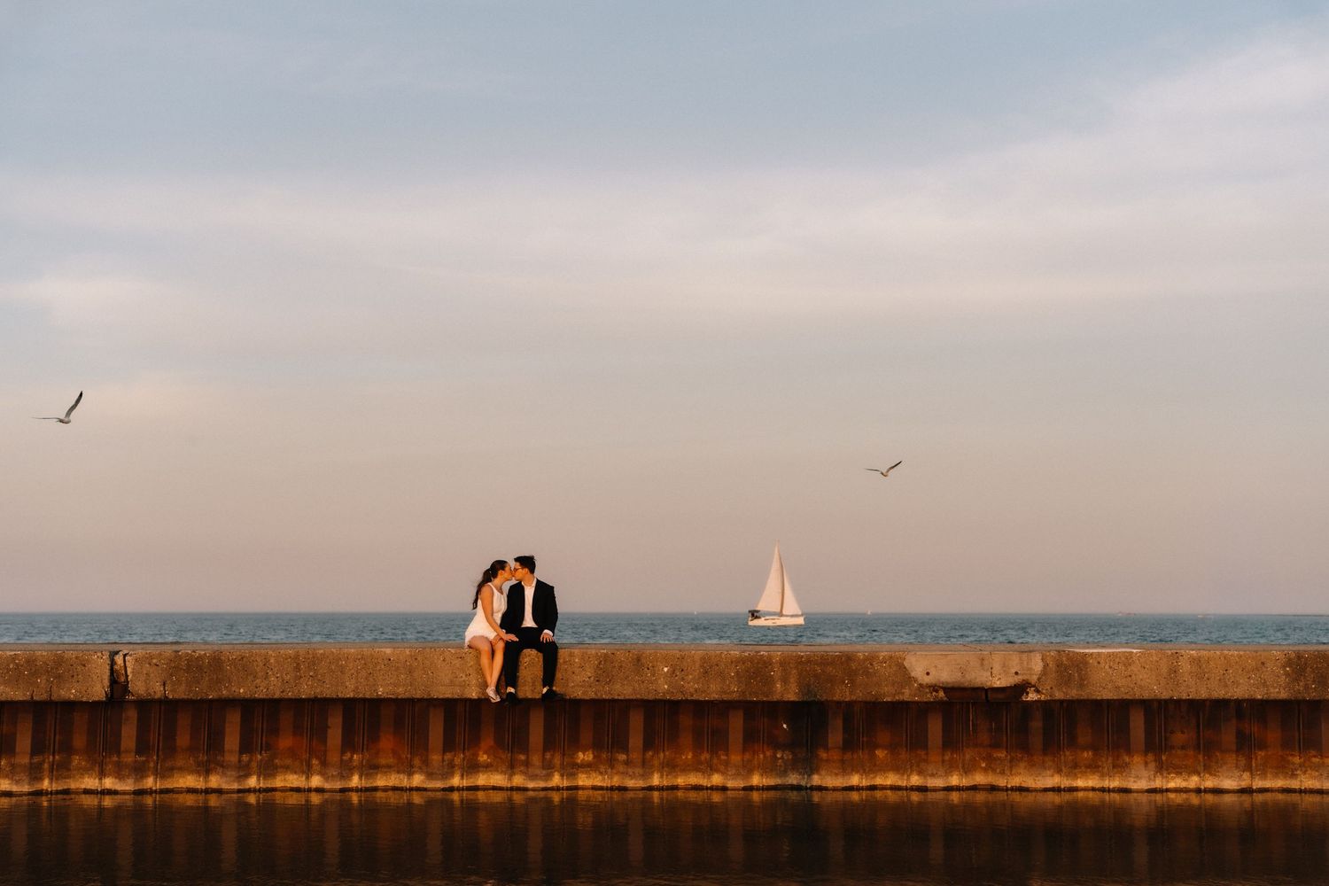 A couple sits close together on a seawall at sunset, with a sailboat and seagulls over the calm lake water in the background.