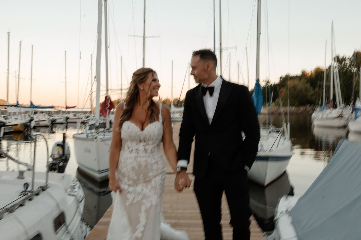 Bride and groom walk hand-in-hand on a marina dock at sunset, surrounded by sailboats.