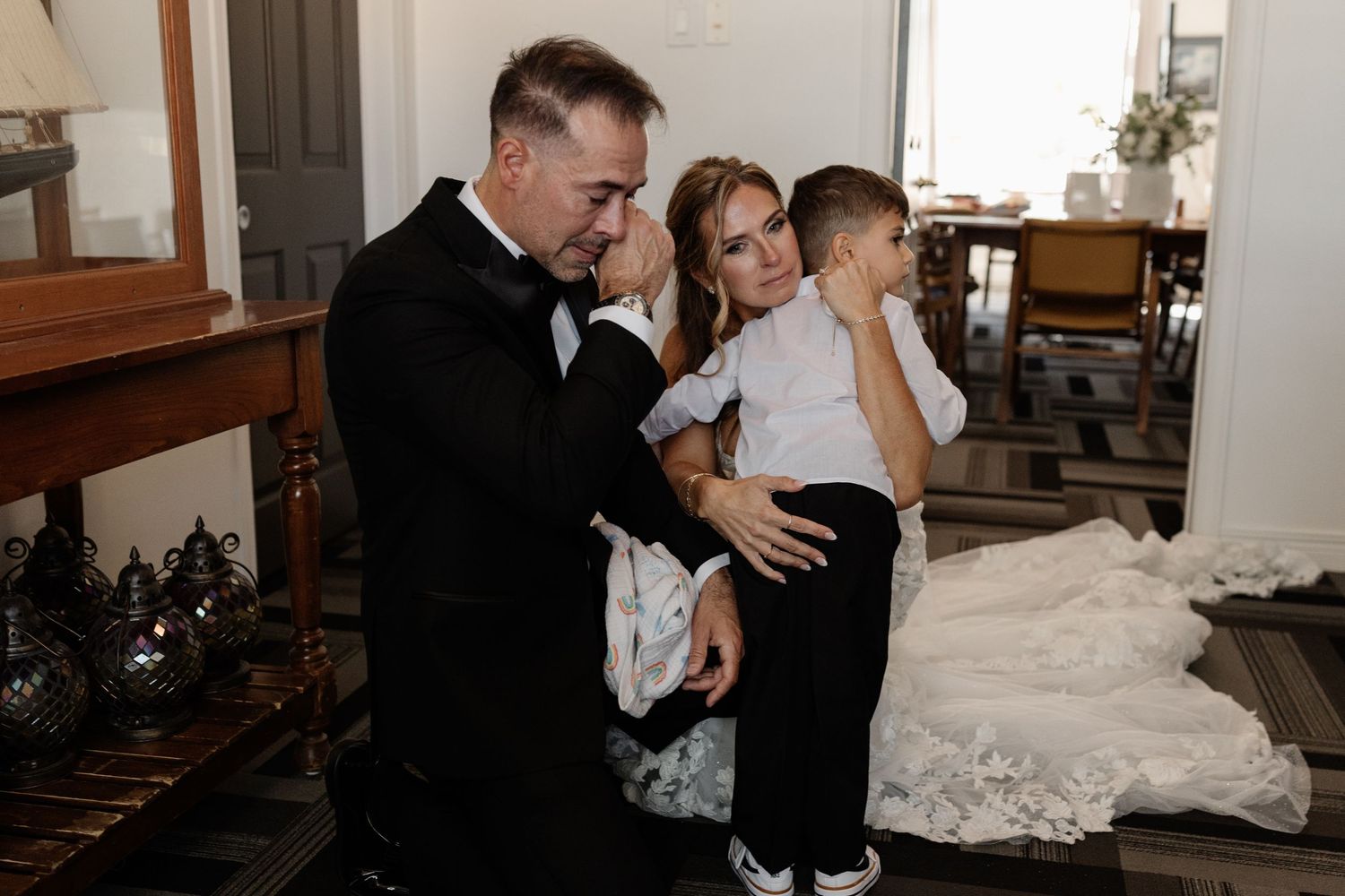 A couple in wedding attire tenderly holds a young child in a warmly lit room.
