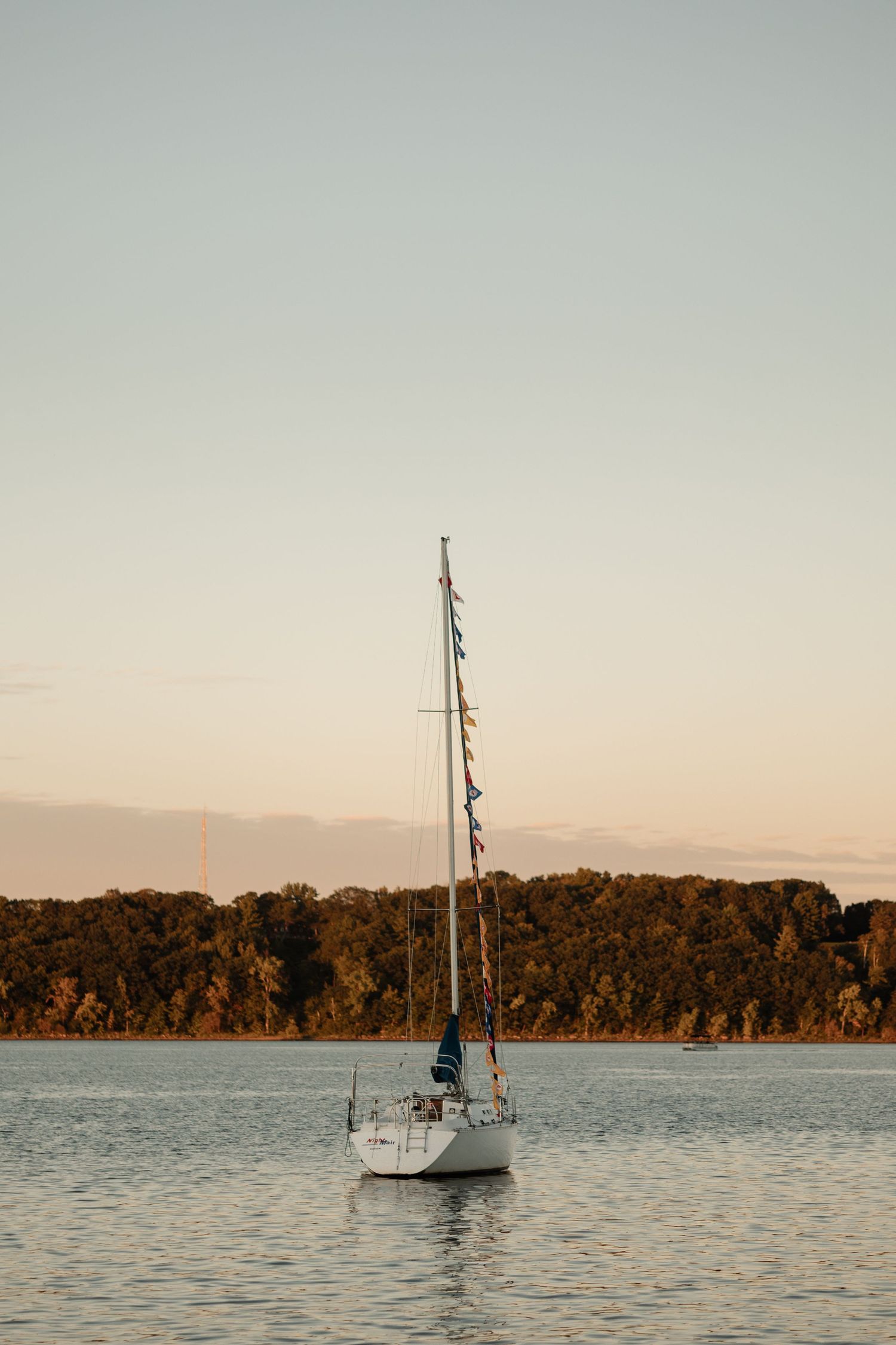 White sailboat anchored on calm lake waters at golden sunset, with lush autumn forest shoreline in background.