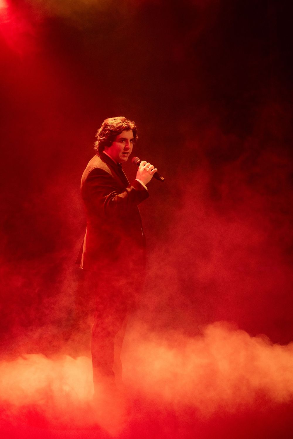 Male singer performing on stage with microphone, surrounded by dramatic red lighting and atmospheric fog effects.