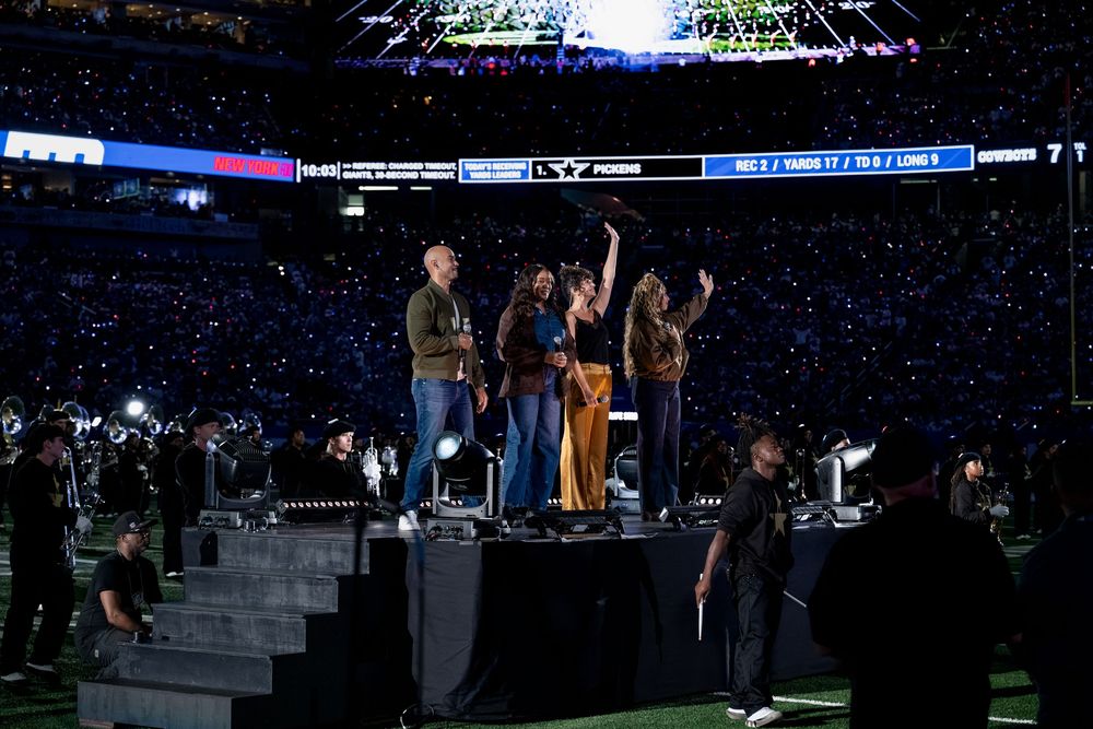 Group of performers on a stage at a large stadium concert event with crowd and scoreboard visible in background.