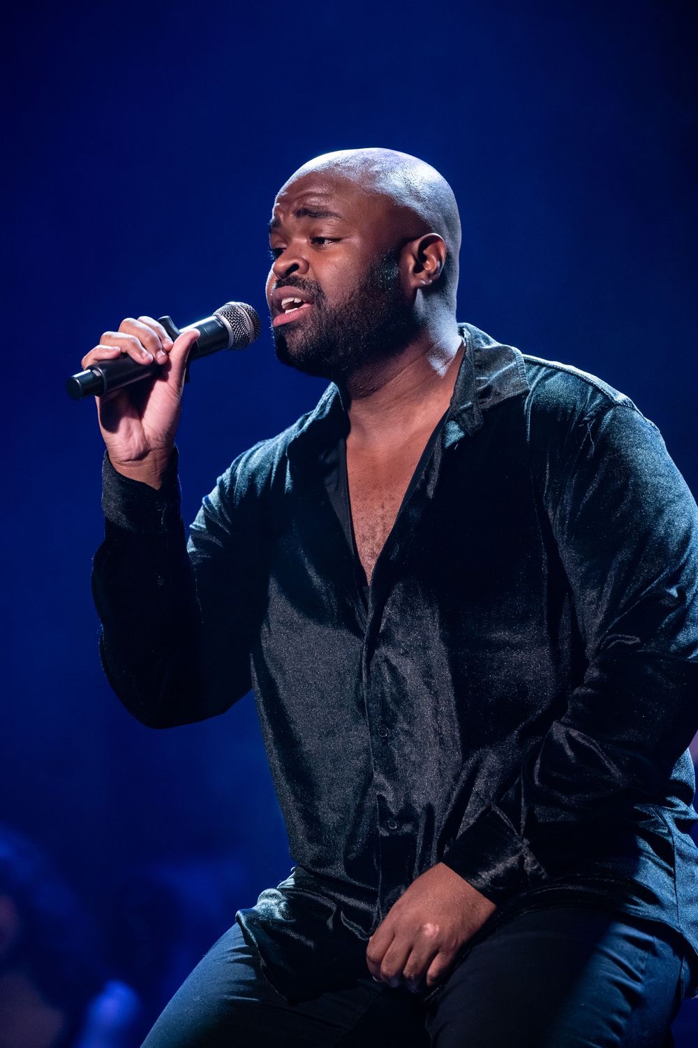 Bald male singer performing on stage with microphone, wearing dark velvet shirt under blue stage lighting.