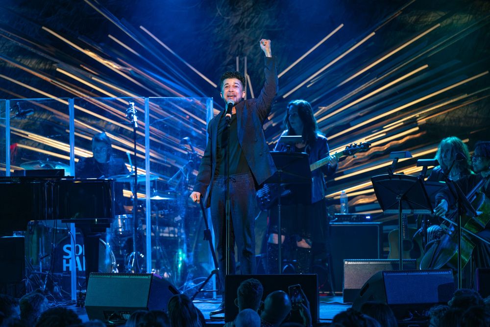 Singer performs on stage with fist raised, dramatic lighting and band backdrop at live concert.