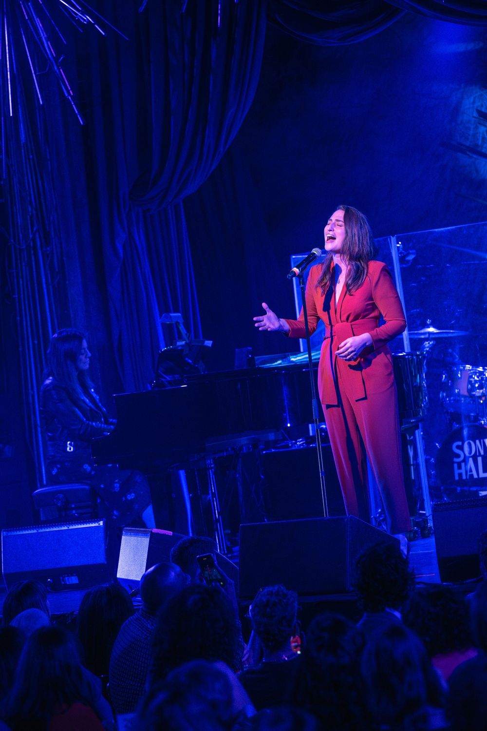 Woman in red suit performing on stage at a live music venue with blue lighting and audience in foreground.