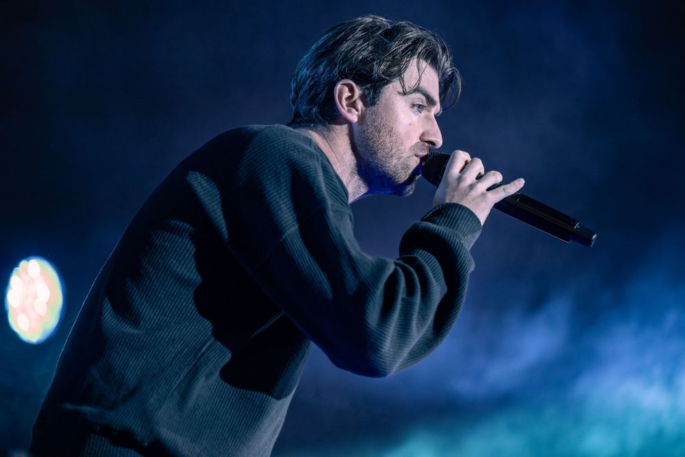 Male singer performing on stage holding microphone in dramatic blue smoky lighting wearing dark sweater.
