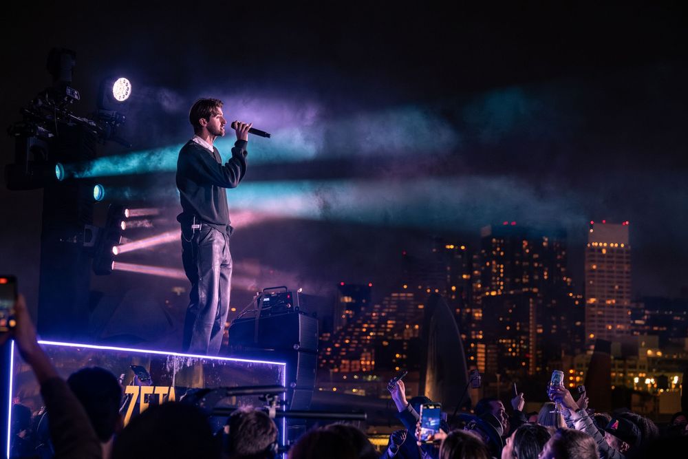 Singer performs on illuminated outdoor stage at night with city skyline and colorful stage lights in background.