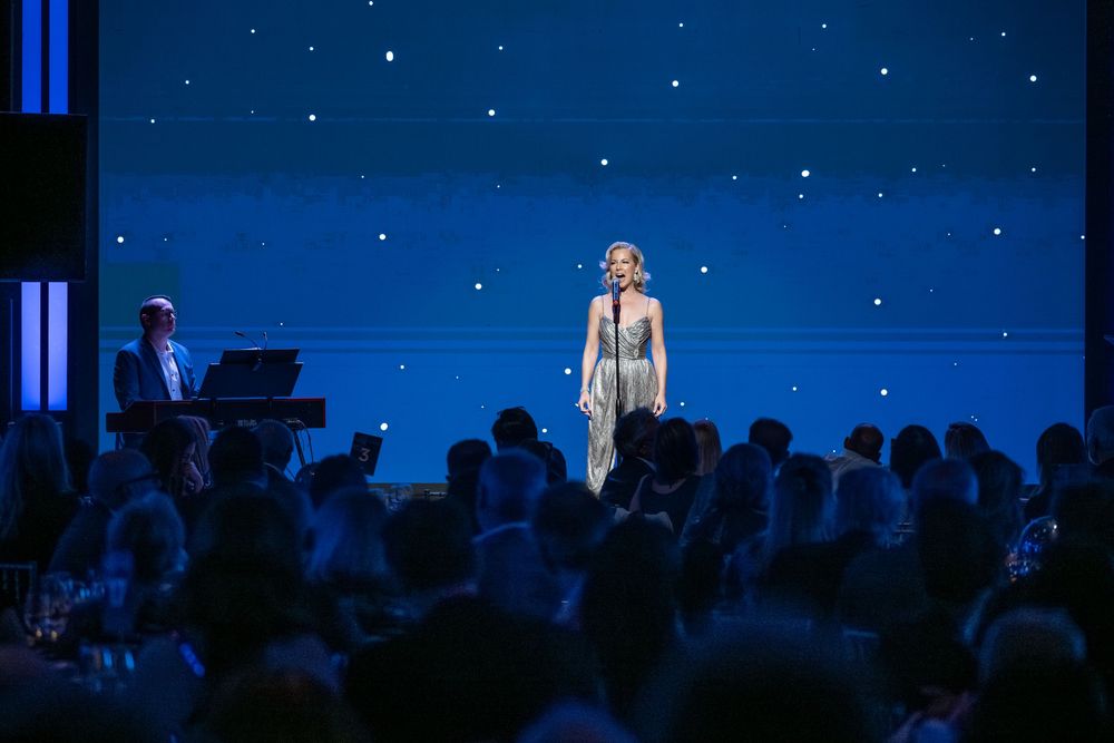 A woman in a silver gown performs on stage before a seated audience under a starry blue backdrop with a pianist.