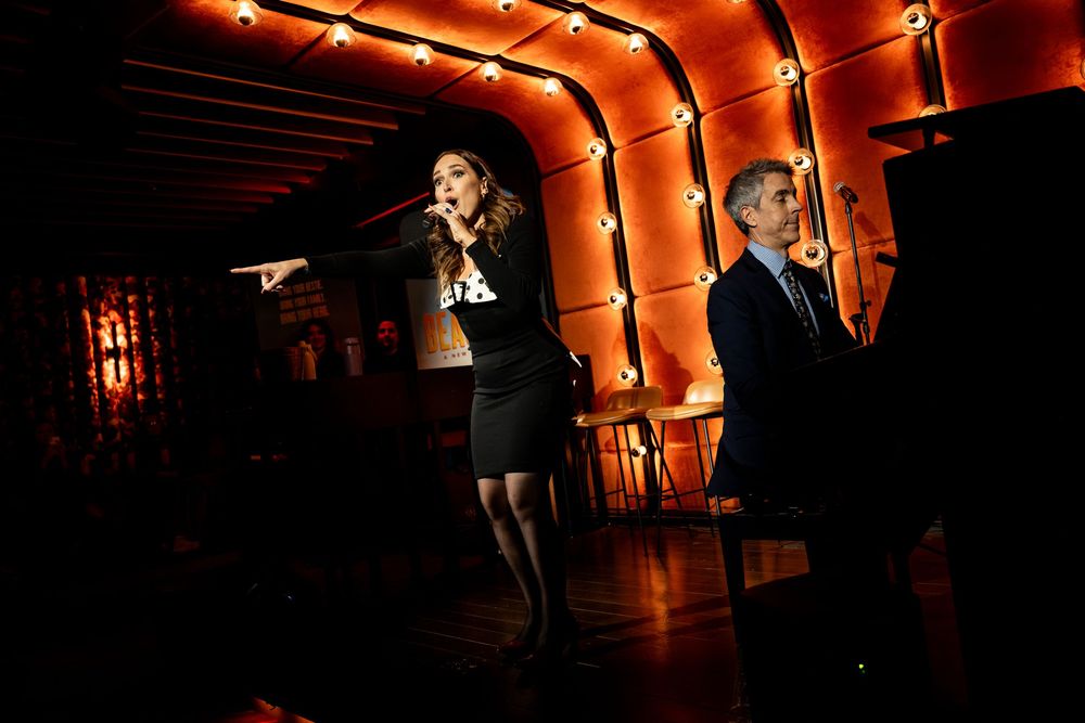 A woman sings into a microphone while a man plays piano on a dimly lit stage with warm orange lighting.