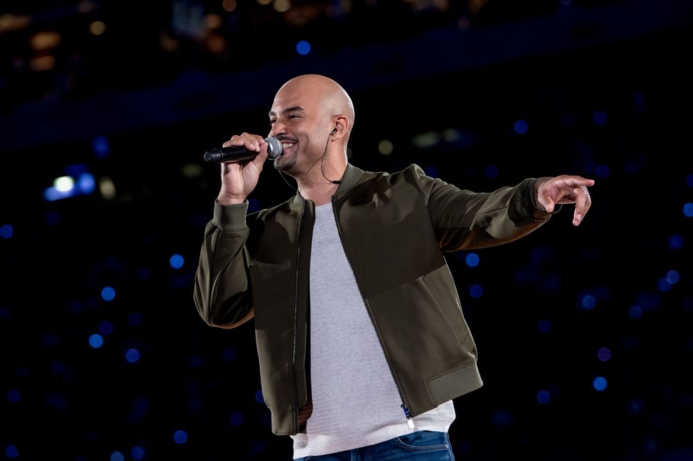 Bald male singer performing on stage, holding microphone and pointing, wearing green bomber jacket and white shirt.