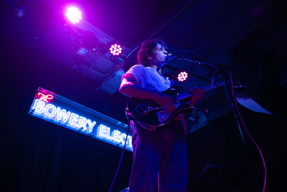 Musician performs on stage at Bowery Electric venue, playing guitar under colorful purple and pink stage lights.