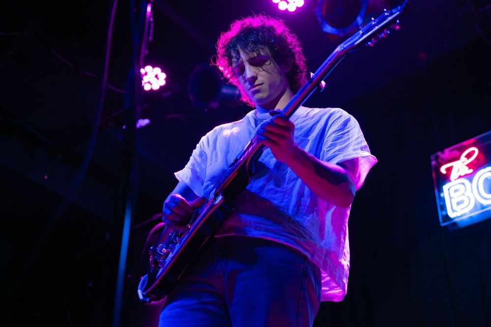 Young male guitarist performing on stage under purple and blue lights at a live music venue.