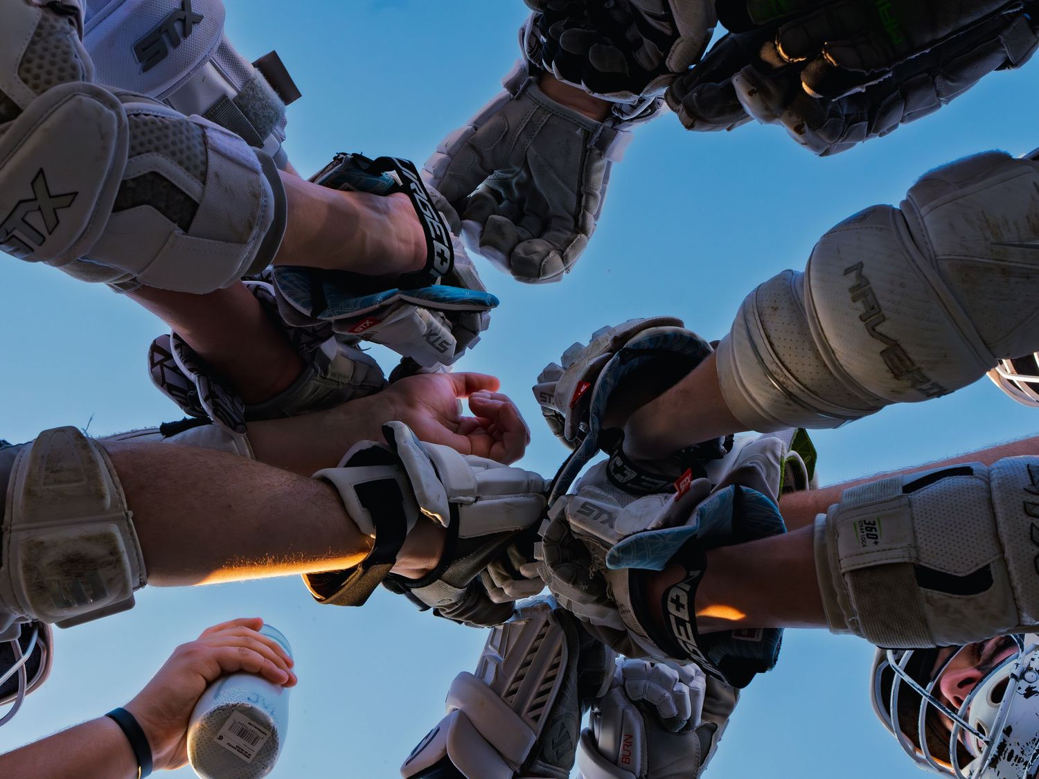 Janesville lacrosse players stack gloved hands together in a team huddle, shot from below against a clear blue sky before the game.