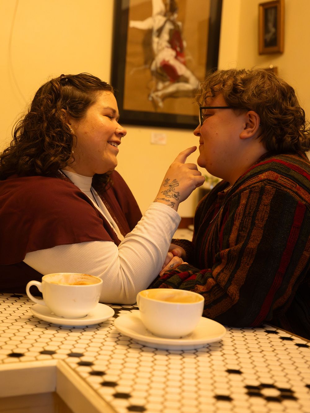 Two people share a playful moment over coffee cups at a cafe table, one touching the other's chin affectionately.