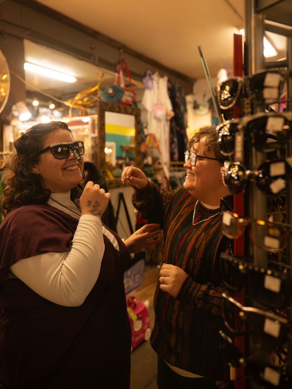 Two women laughing and trying on sunglasses in a colorful accessory shop.