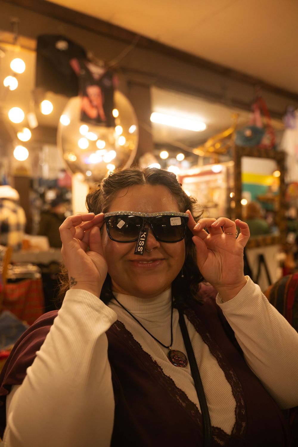 Woman trying on oversized Guess sunglasses smiling in a vintage boutique shop with warm bokeh lights.