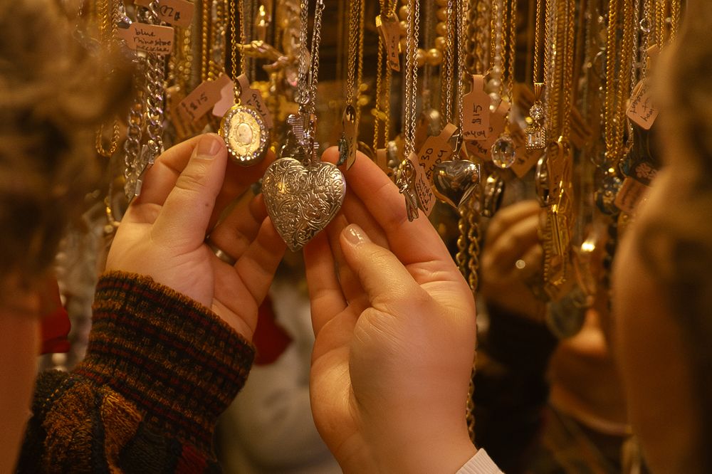 Hands holding a silver heart locket among rows of gold necklaces at a jewelry market stall.