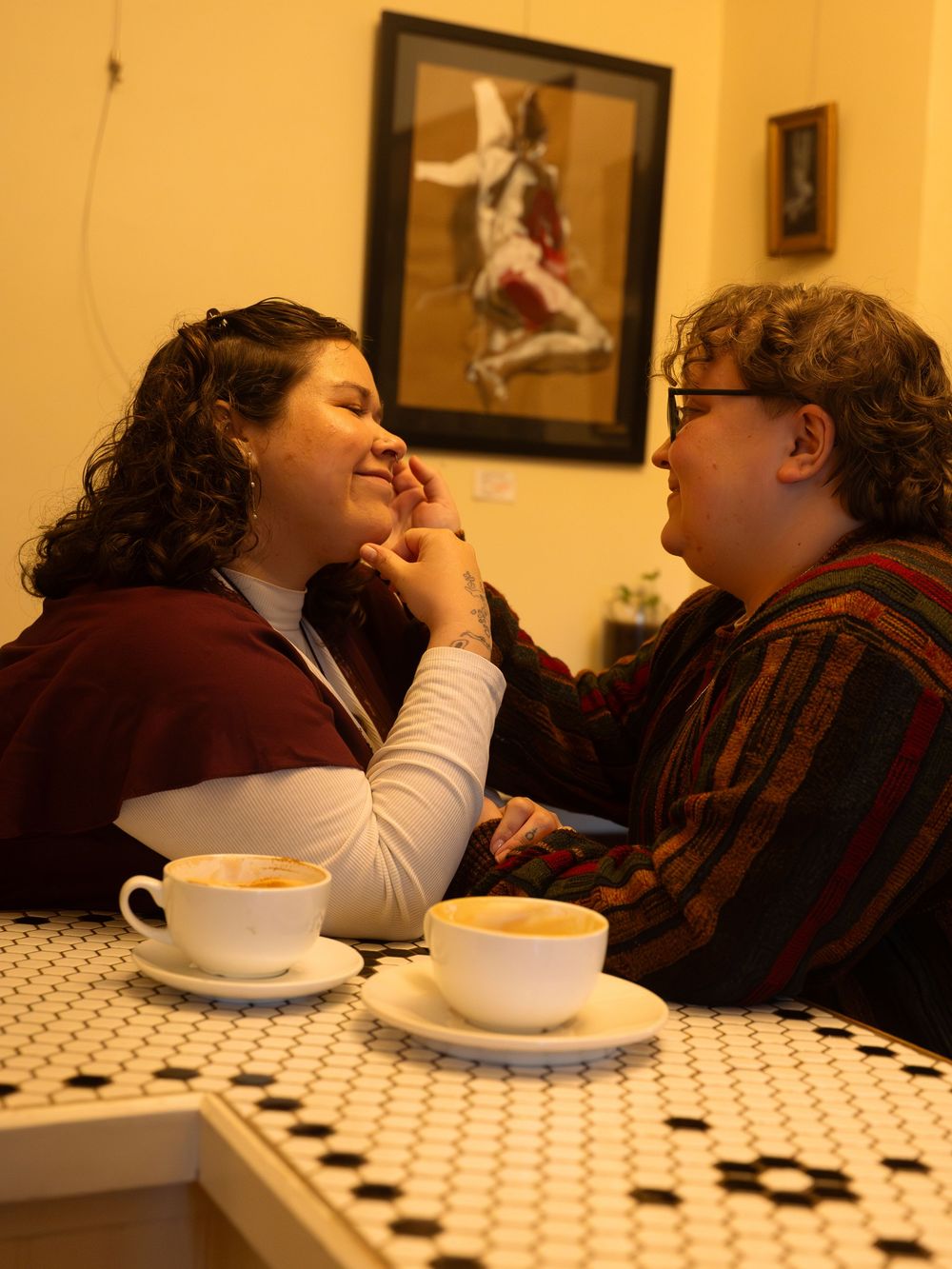 Two people share a romantic moment over cappuccinos at a cafe table with a mosaic top.