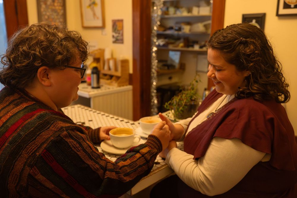 Two women share a warm moment over coffee in a cozy cafe decorated with Christmas lights.