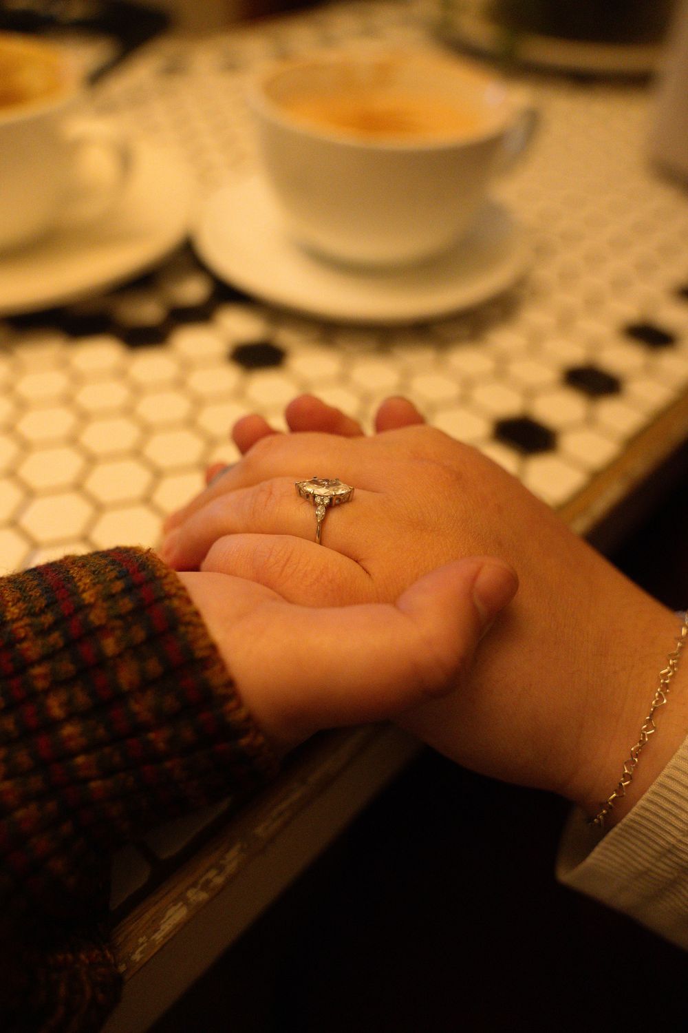 Two hands holding on a mosaic table, one wearing a diamond engagement ring, with coffee cups in the background.