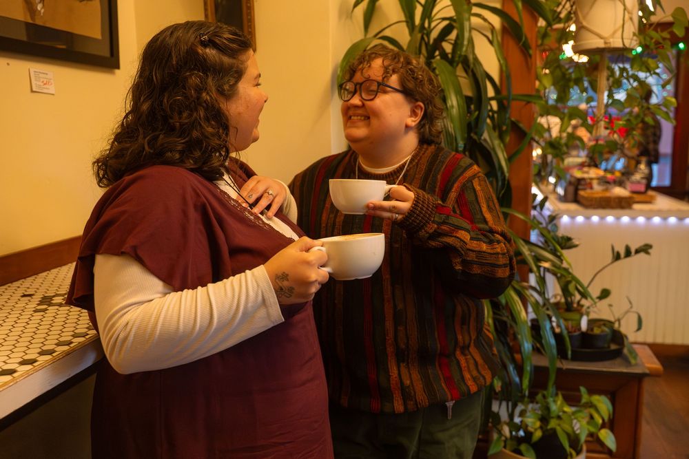 Two women holding white mugs chat warmly indoors near lush green plants and cozy cafe decor.