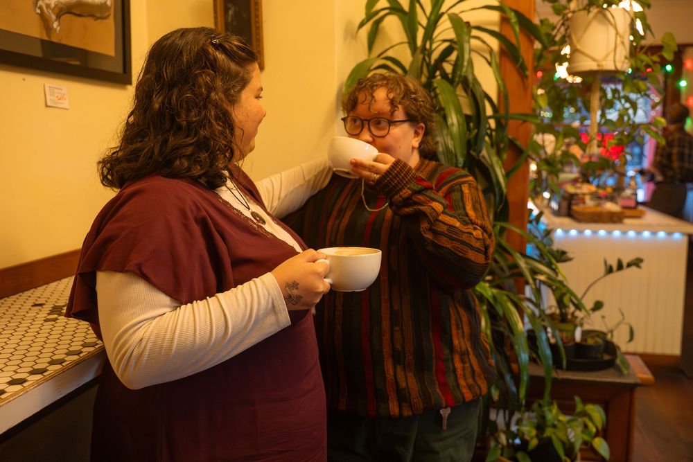 Two women share a warm moment over coffee cups near a lush indoor plant in a cozy cafe.
