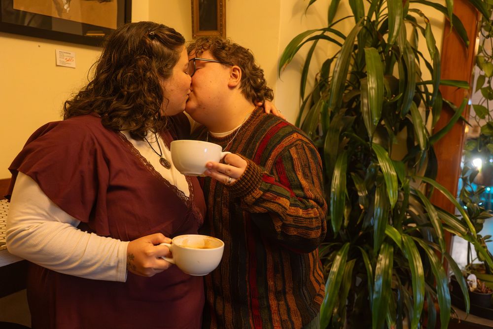 Two people sharing a kiss while holding coffee cups near a tropical plant indoors.