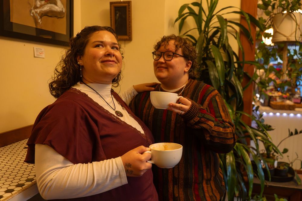 Two women smiling and holding coffee cups indoors near a lush green plant in warm ambient lighting.