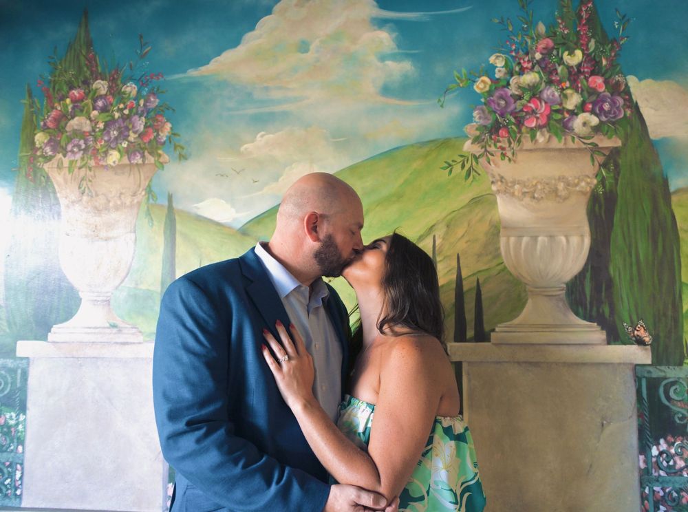 Couple sharing a romantic kiss in front of a colorful painted mural with floral urns and Tuscan landscape backdrop.