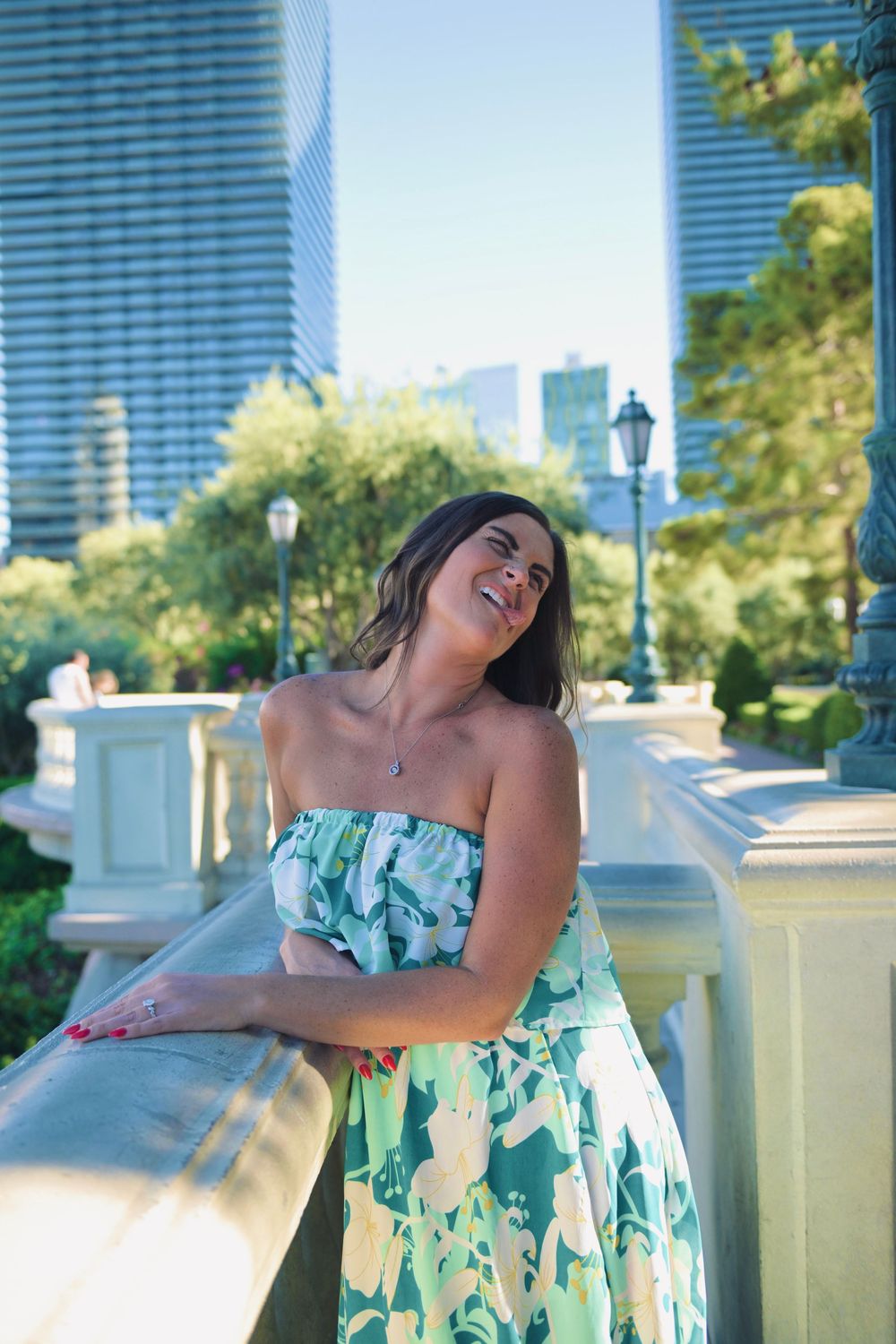 Woman in green floral strapless dress smiling outdoors with city skyscrapers and trees in background.