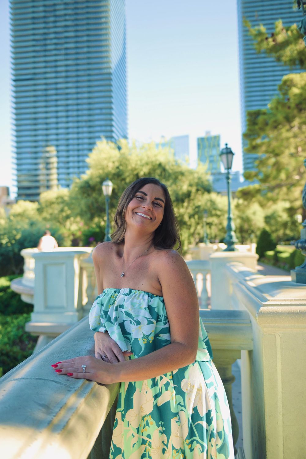 Woman in green floral strapless dress smiling on balcony with city skyscrapers in background.