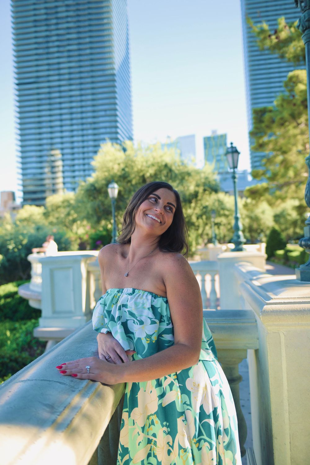 Smiling woman in green floral strapless dress leaning on balcony railing with city skyscrapers in background.