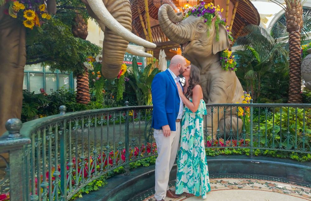 Couple sharing a kiss near elephant statue in lush botanical garden with colorful floral decorations.