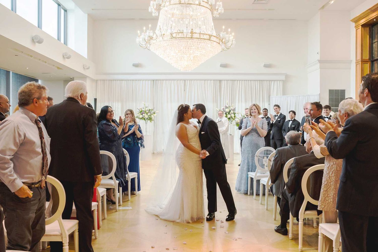 Bride and groom share first kiss at elegant indoor wedding ceremony with chandelier and applauding guests at view venue