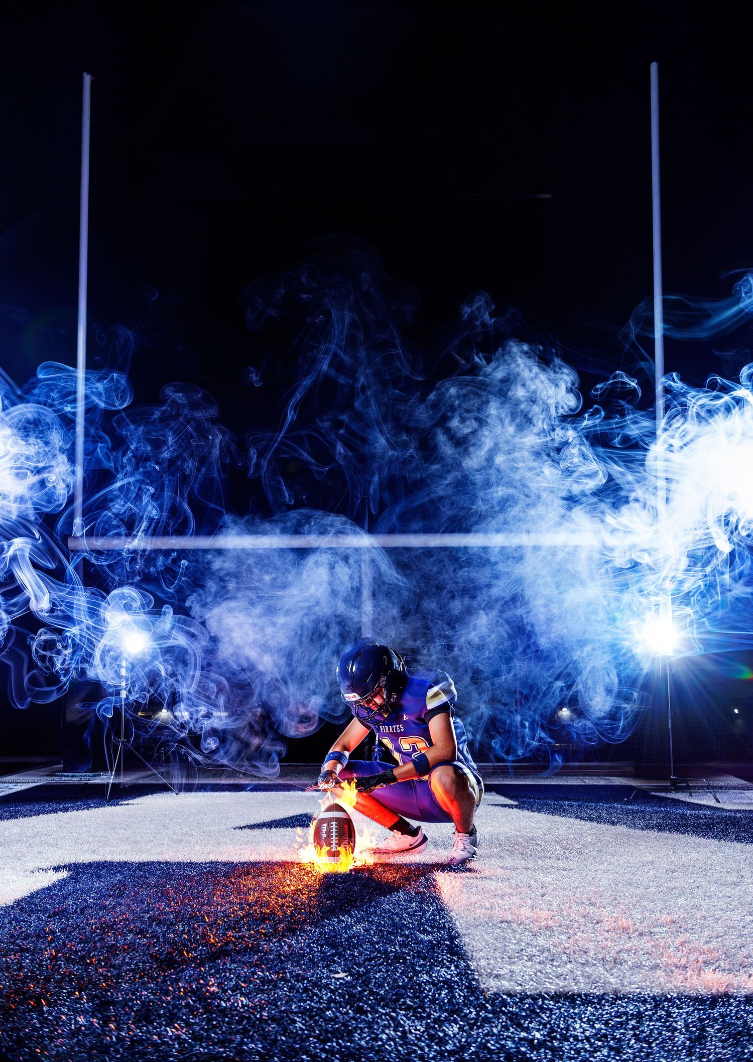 Football player kneeling on field at night surrounded by dramatic blue smoke with glowing light and goalposts in background.