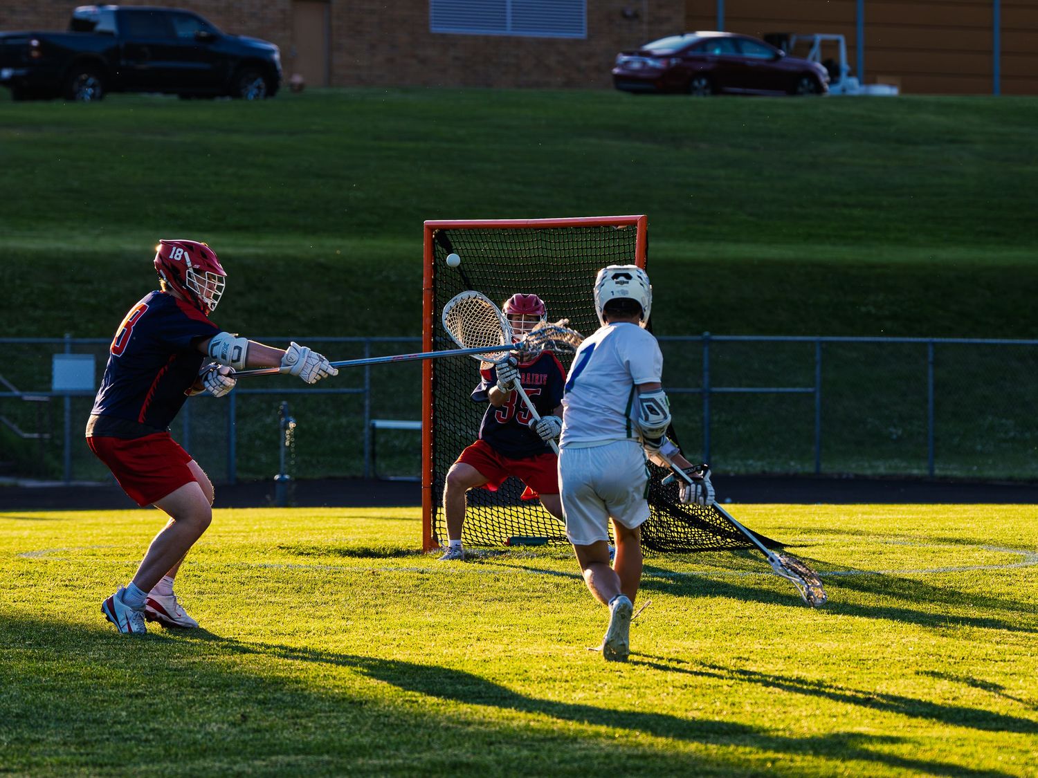 Janesville lacrosse player in white fires a shot as the ball sails toward the net past a red-jersey Prairie goalie in golden evening sun.