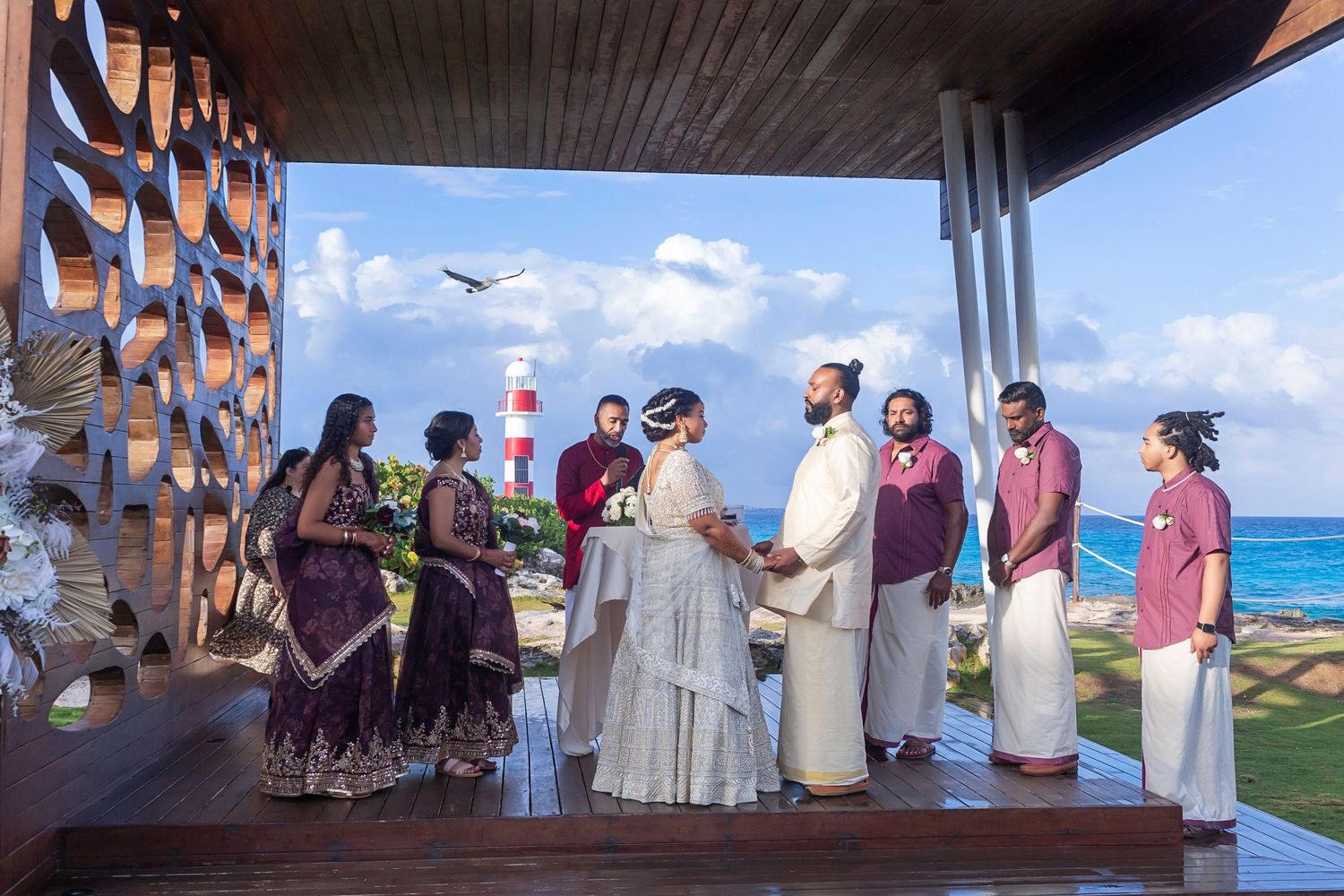 South Asian couple in traditional gold wedding attire posing on rocky shoreline with lighthouse in background.