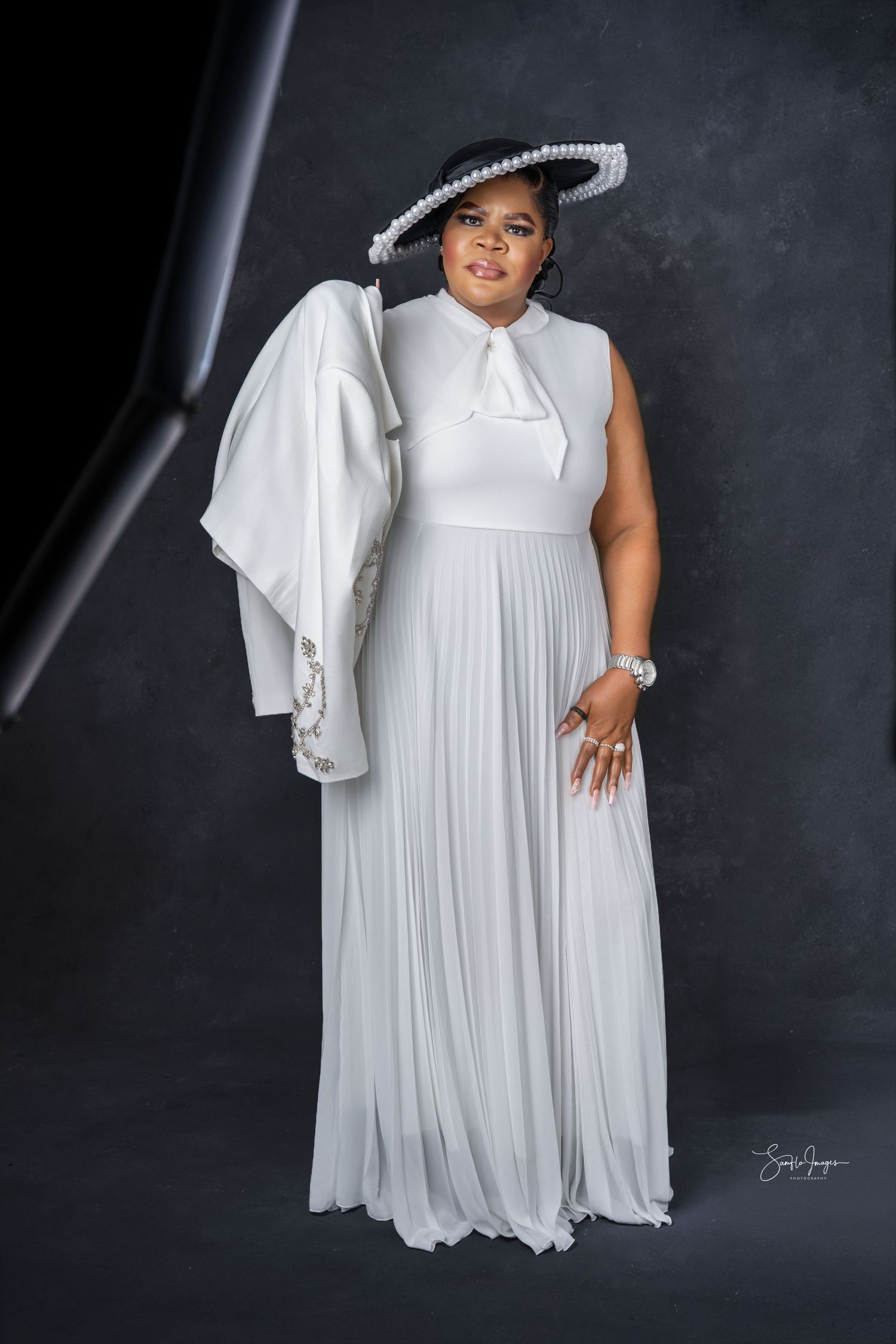 Woman in elegant white pleated gown and black pearl-trimmed hat poses gracefully holding a white jacket.