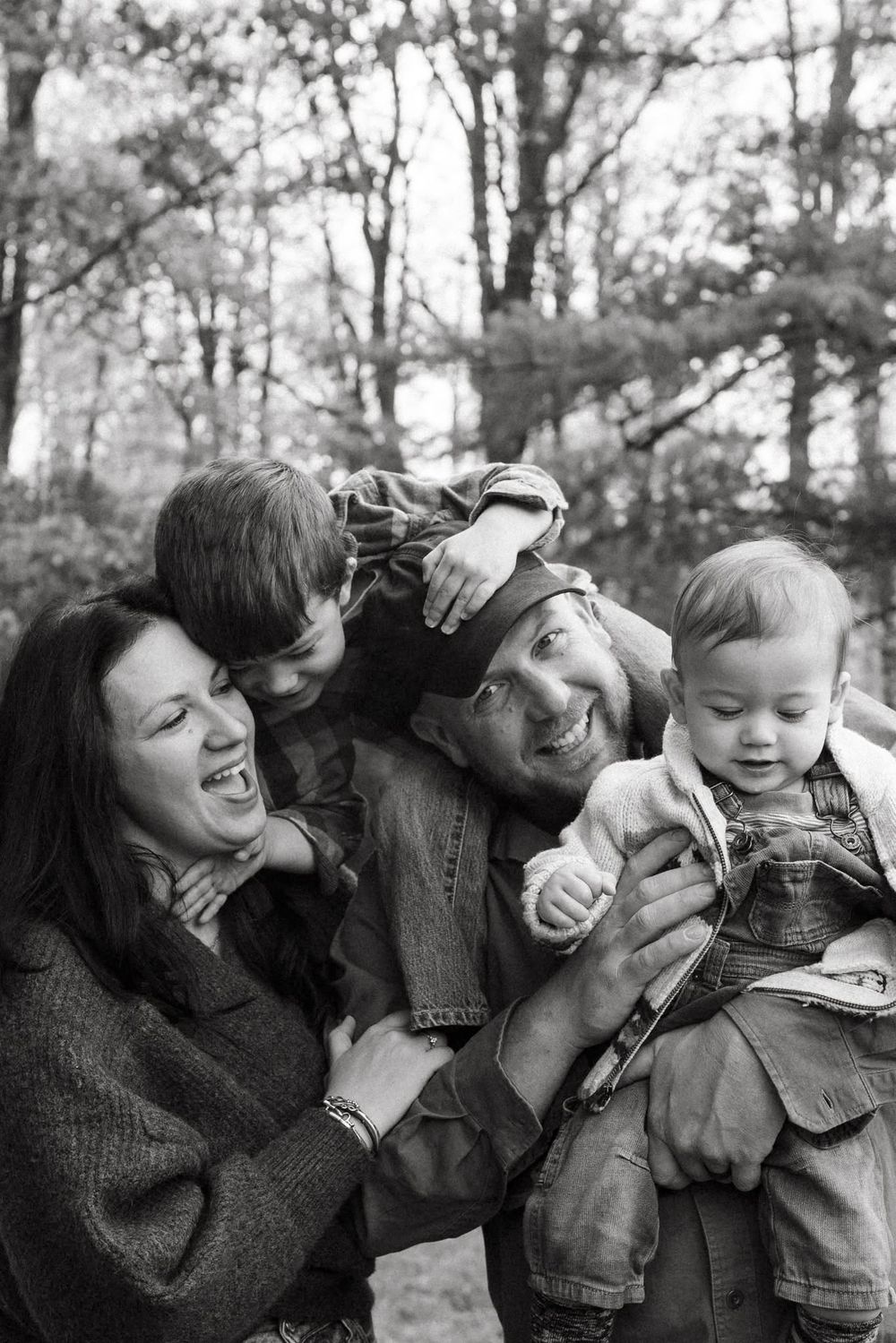 Black and white family portrait of parents with two children laughing outdoors in a wooded setting.