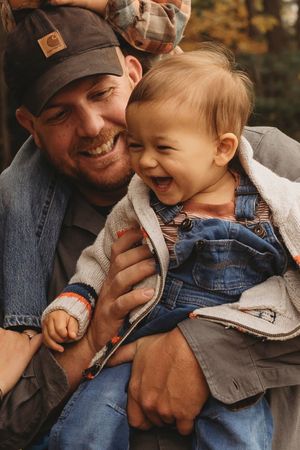 A smiling man in a Carhartt cap holds a laughing toddler in denim overalls outdoors in autumn.