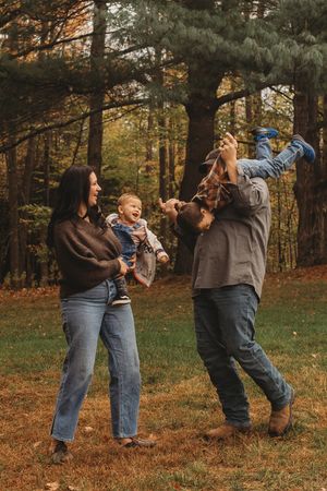 Family of four playing outdoors in autumn forest, parents laughing while lifting children among colorful fall foliage.