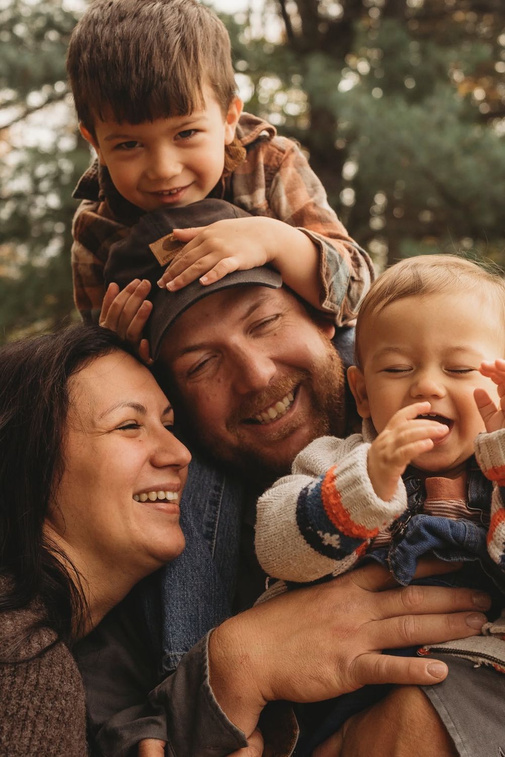 Happy family of four laughing outdoors, parents holding two young boys, surrounded by green trees in warm autumn light.