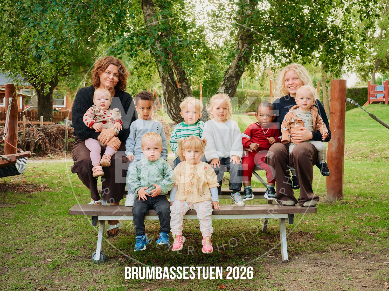 Group photo of two women with several young children on a bench outdoors, labeled Brumbassestuen 2026.