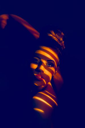 Woman with golden light stripes casting dramatic shadow patterns across her face in dark blue studio portrait.