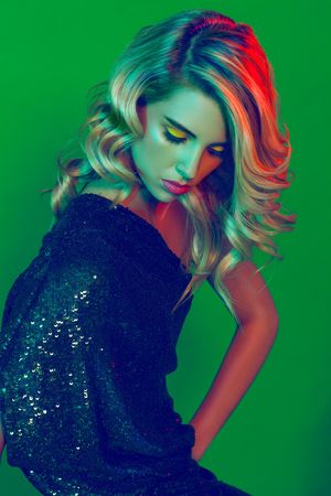 Blonde woman in black sequin dress with bold yellow eye makeup posed under vibrant green and red studio lighting.